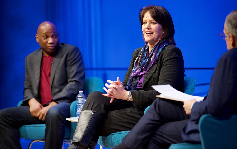 Three public program participants sit on a blue-lit auditorium stage. To the far left, a man in a gray blazer, red sweater, and jeans looks over to a woman in the center, who addresses an unseen audience. The moderator, partially cut off in the photograph, sits with his back to the camera and holds a clipboard in his lap.