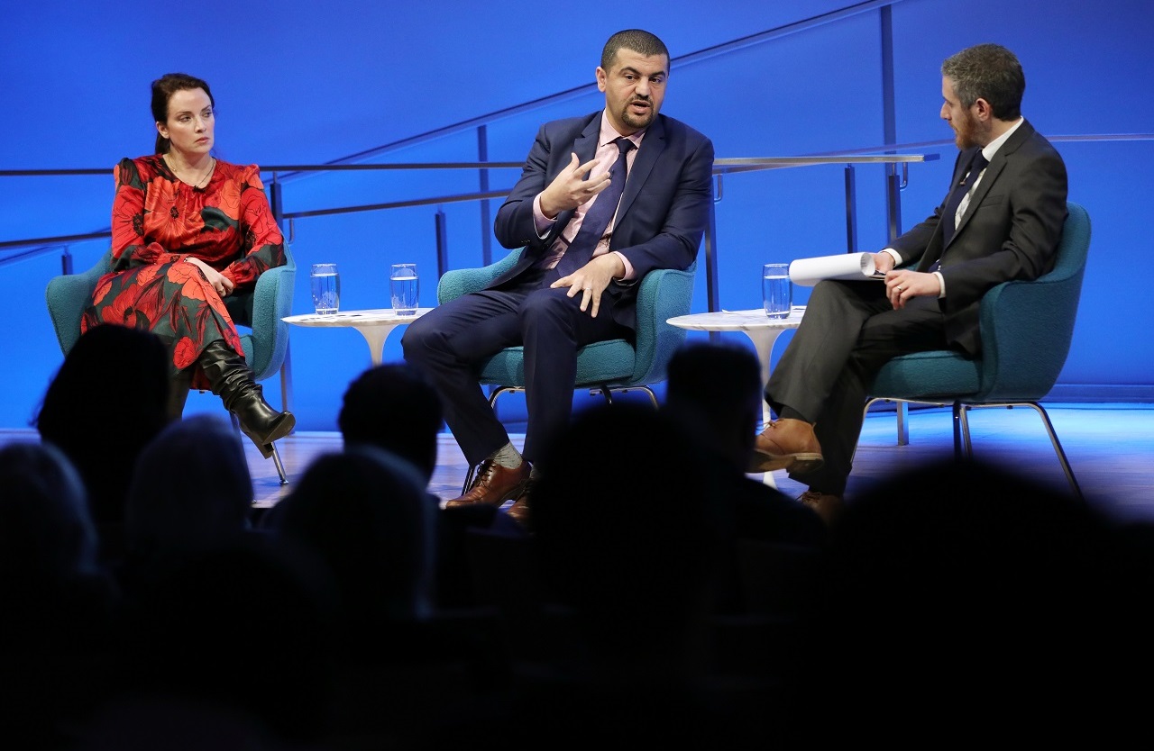 Journalist Hassan Hassan holds up his right hand as he speaks onstage and looks towards the audience at the Museum Auditorium. American Enterprise Institute scholar Karen E. Young listens to Hassan’s right. Moderator Noah Rauch listens to Hassan’s right as he holds a clipboard.