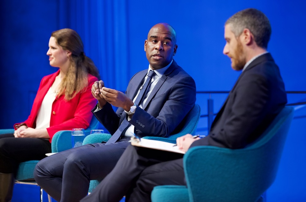 Three public program participants sit on a blue-lit auditorium stage in this close-cropped photograph. On the far left, a woman in a red blazer looks off toward an unseen audience. In the center, a man gestures with his hands and looks in the direction of the moderator. The moderator sits with a clipboard in his lap and looks down while listening.