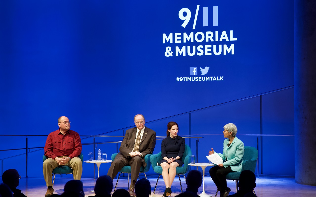 A moderator leading the discussion in a public program at the Museum Auditorium gestures with her right hand and holds a piece of paper with her left. She is looking at three guests seated to her right. They are artist Melissa Cacciola, Local 40 Business Manager Robert Walsh, and Kahnawake Council Chief Lindsay LeBorgne. The three of them are listening with their hands in their laps. Audience members are silhouetted in the foreground by the lights onstage.