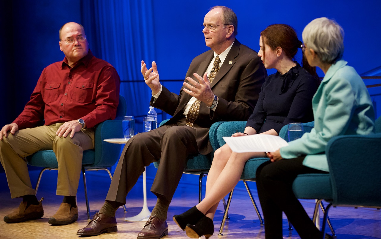 Local 40 Business Manager Robert Walsh speaks while seated onstage during a public program at the Museum Auditorium. He is holding up both hands as he looks out towards the audience, who are not visible. To his right is Kahnawake Council Chief Lindsay LeBorgne. To his left is artist Melissa Cacciola and the female moderator of the discussion. The moderator is in the foreground and is out of focus.