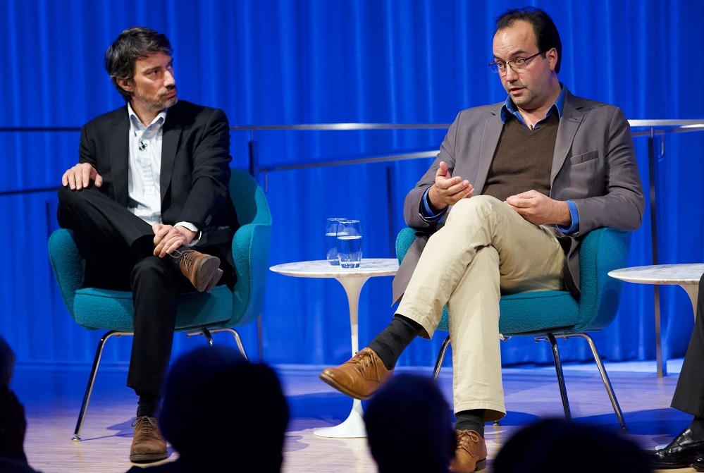 Two men in sportcoats sit on an auditorium stage.