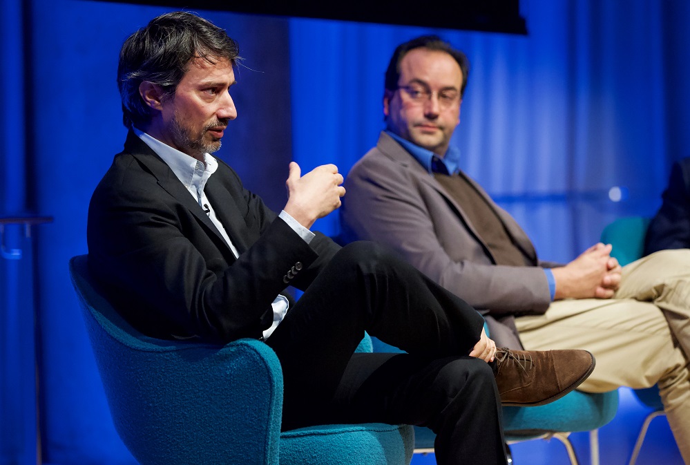 Two men in sportcoats sit on an auditorium stage.