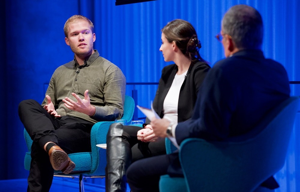 Two men and a woman sit on a blue-lit auditorium stage. A man to the far left speaks while the other two look on.