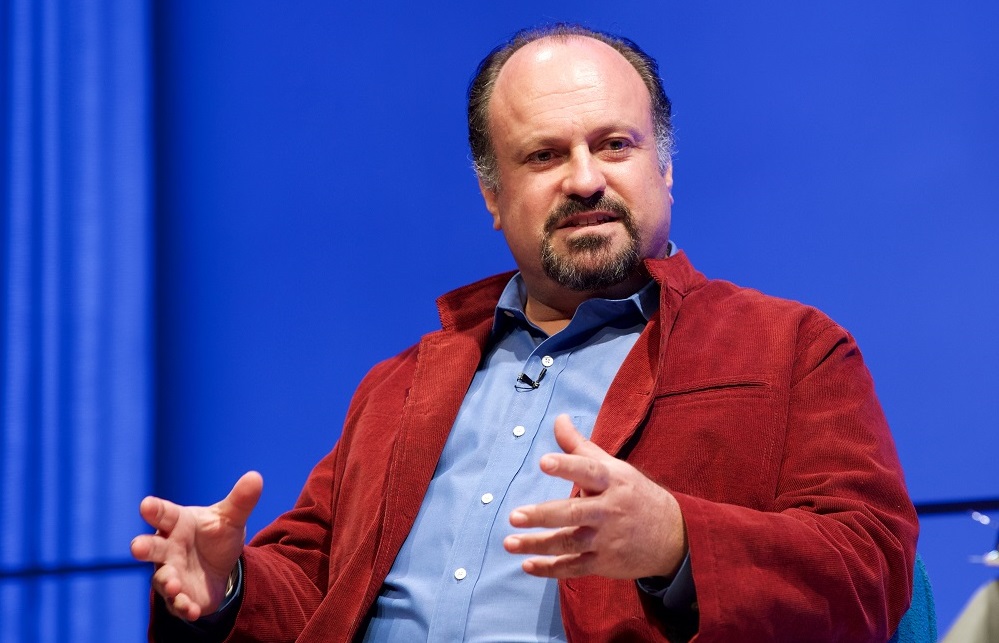 In this close-cropped photograph, a man with a red jacket gestures with his hands on a blue-lit auditorium stage.