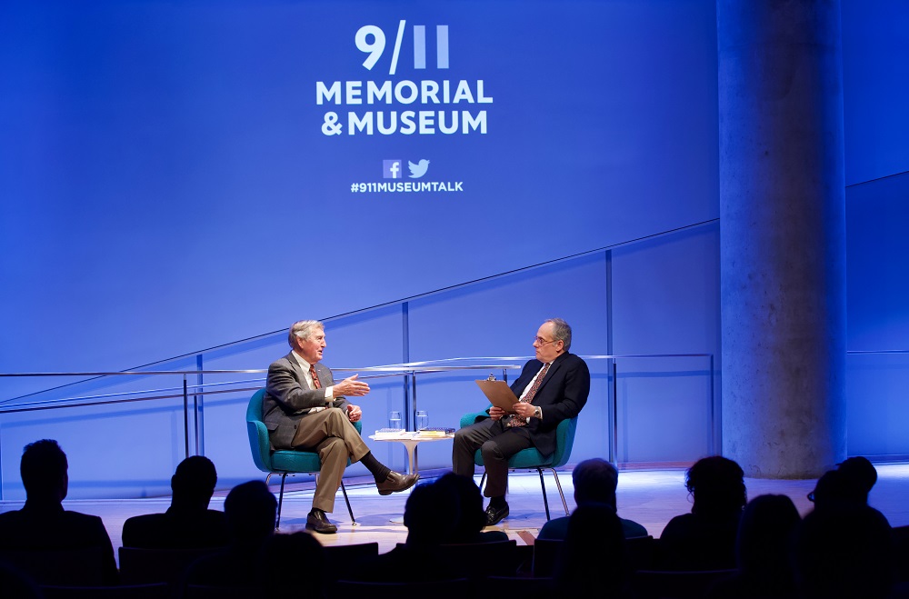 In this wide-angle photograph, two men sit on a blue-lit auditorium stage. The heads of the audience members appear in silhouette in the foreground.