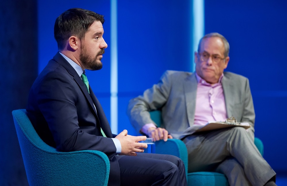 Two men sit on a blue-lit auditorium stage, one in profile speaks while the other looks on.