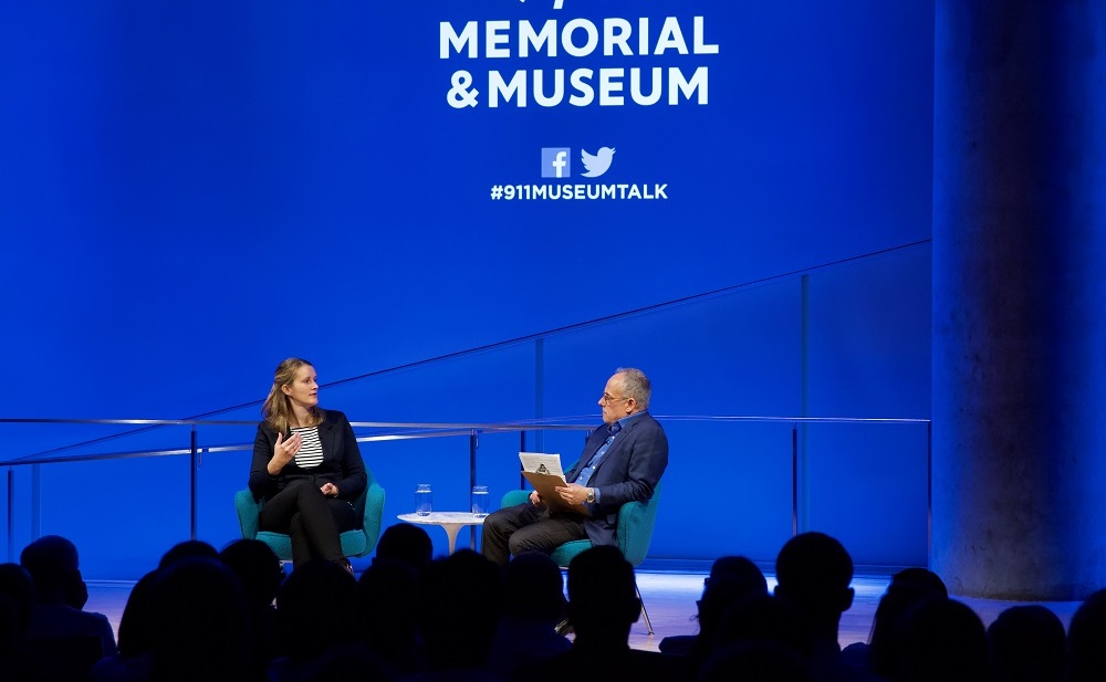 In this wide-angle photograph, a man and a woman sit in conversation on a blue-lit auditorium stage. The heads of audience members appear in silhouette in the foreground.