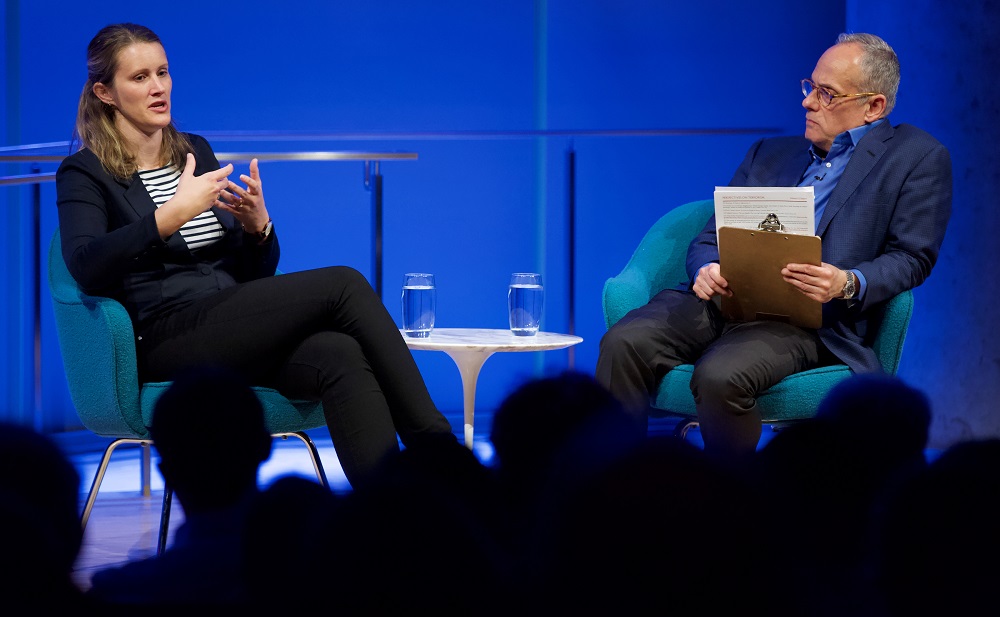 A female speaker gestures with her hands on a blue-lit auditorium stage while the male moderator looks on.