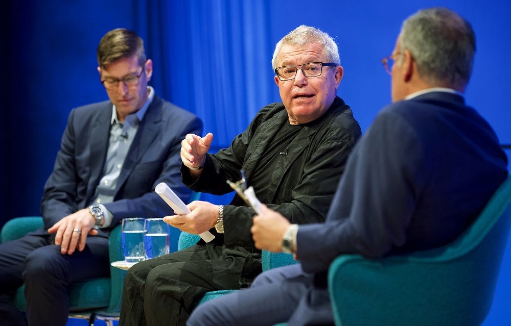 Three men sit on a blue-lit auditorium stage. The man in the center addresses the man closest to the camera while the other man looks down into his lap.