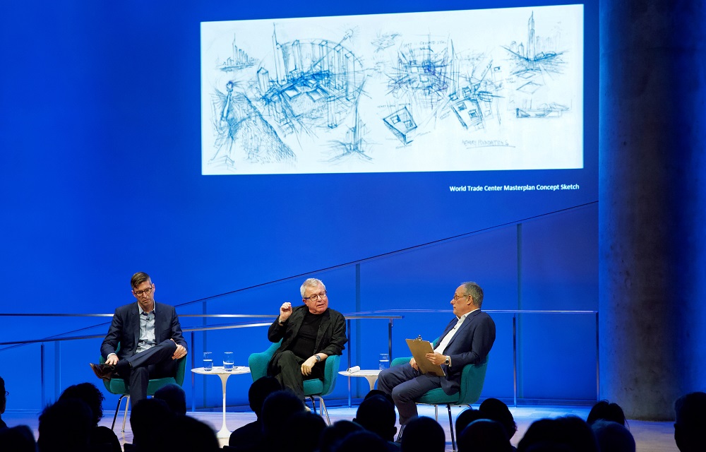 In this wide-angle photograph, three men sit on a blue-lit auditorium stage with an architectural drawing projected onto the wall behind them. The heads of the audience members appear in silhouette in the foreground.