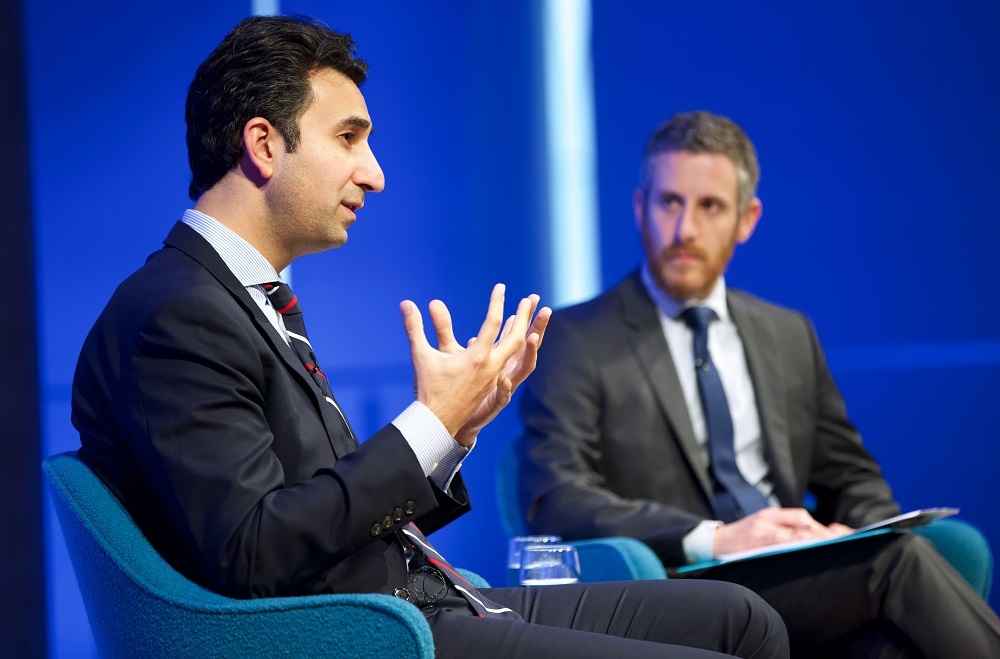 Two men in suits, a public program participant and a moderator, sit on a blue-lit auditorium stage. The program participant appears in profile, gesturing with his hands while speaking out to an unseen audience. The moderator looks on.