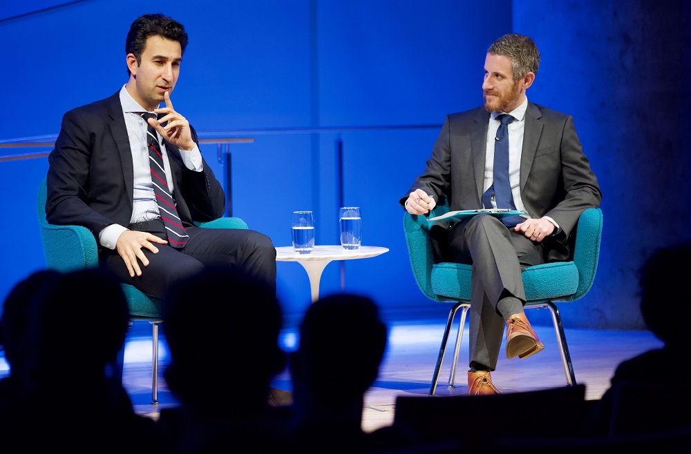 The program participant holds a finger to his lips in thought. The moderator smiles slightly. The heads of the audience members appear in silhouette.