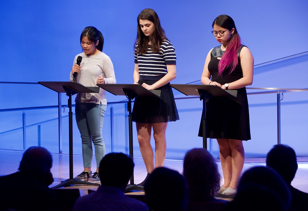 Three people read from podiums on a blue-lit auditorium stage while the heads of audience members appear in silhouette in the foreground.