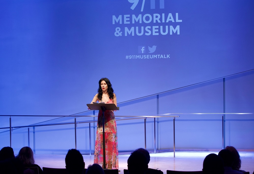 In this wide-angle photograph, one woman reads from a podium a blue-lit auditorium stage while the heads of audience members appear in silhouette in the foreground.