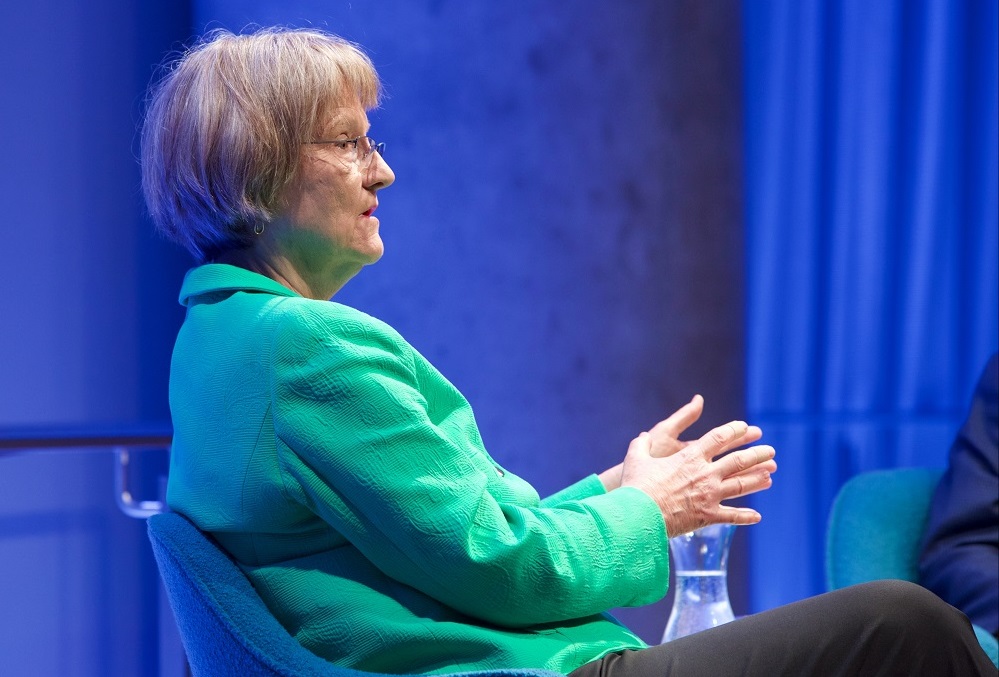 In this close-cropped photograph, a woman in a green suit jacket gestures with her hands while looking out onto an unseen audience.