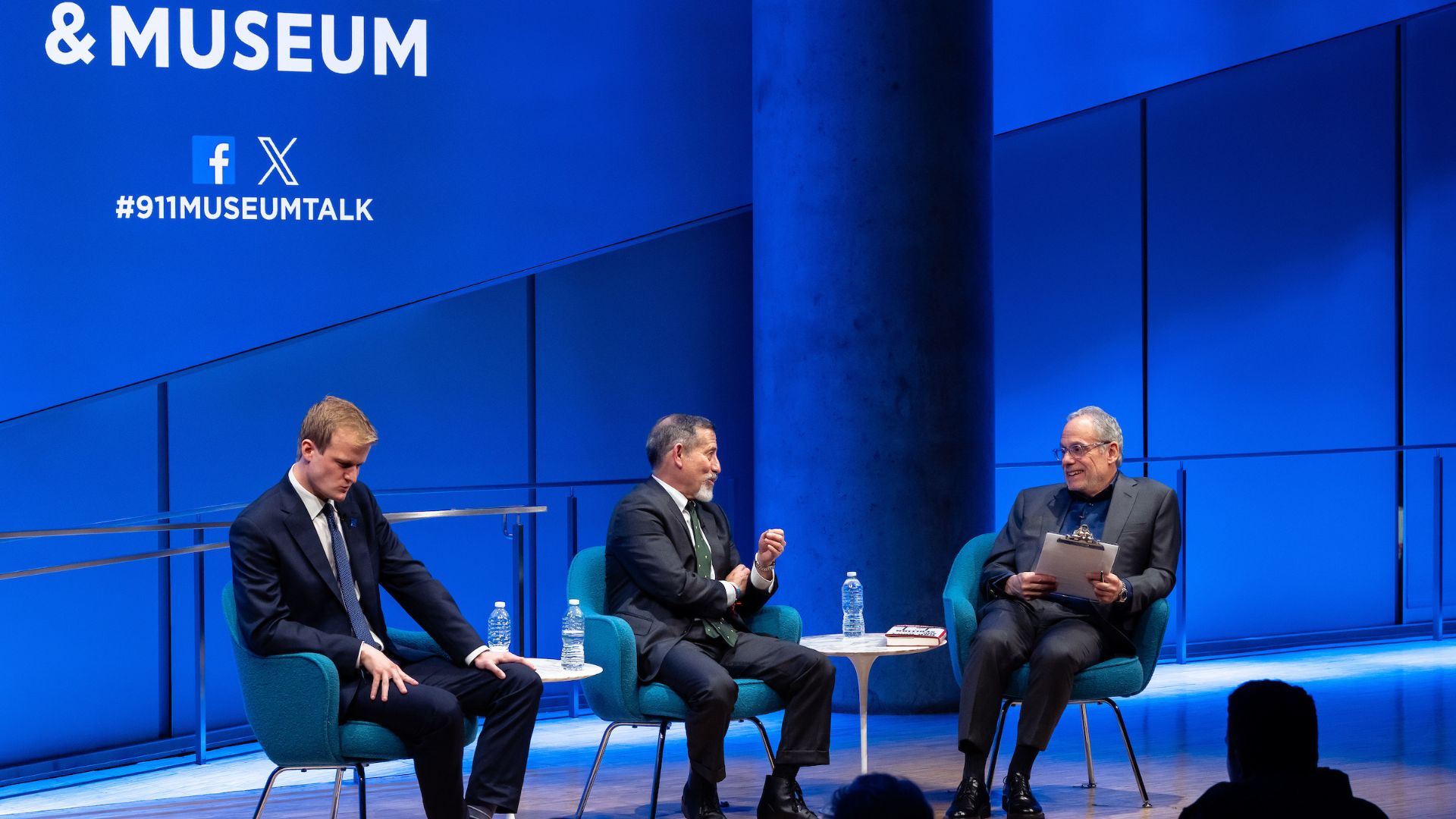 Three men seated on stage, in conversation, in front of a bright blue curtain. A portion of the 9/11 Memorial & Museum logo is visible. 