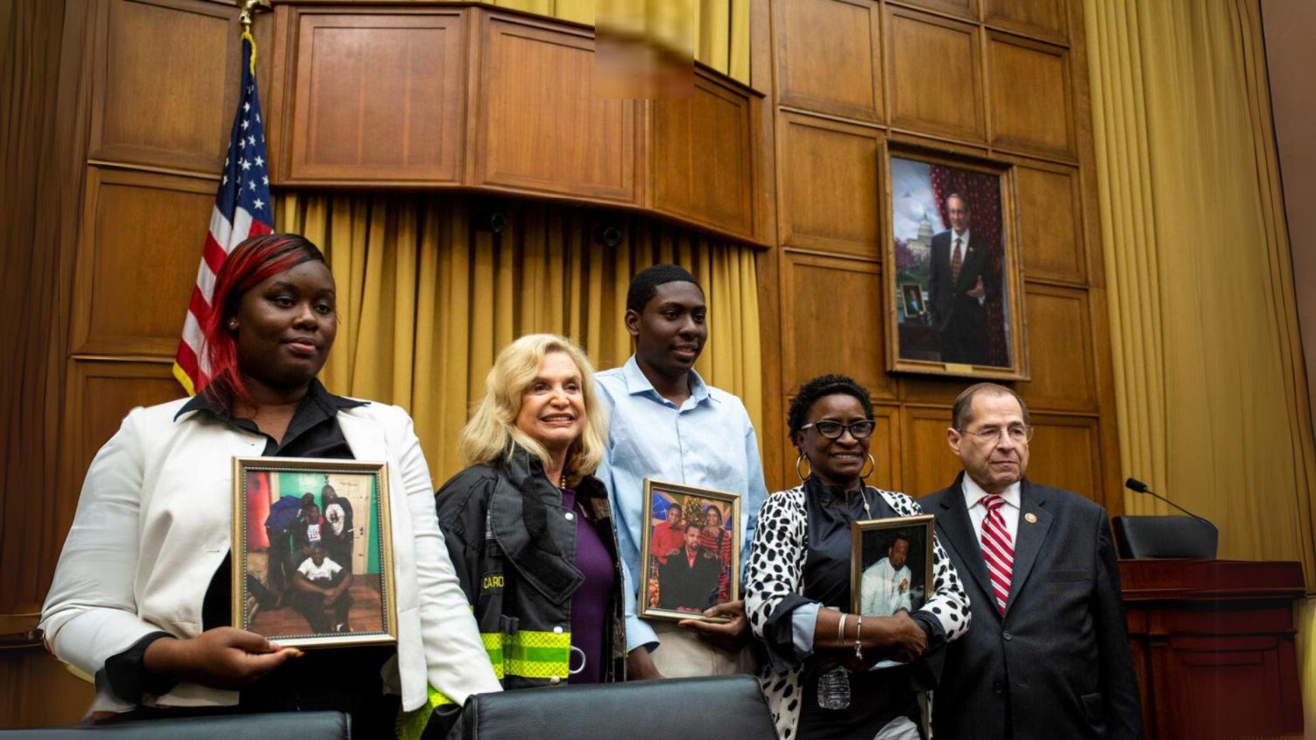 Five people - three of whom hold framed photographs - stand in front of a wood paneled, mustard-color curtained wall. An American flag is visible on the left.