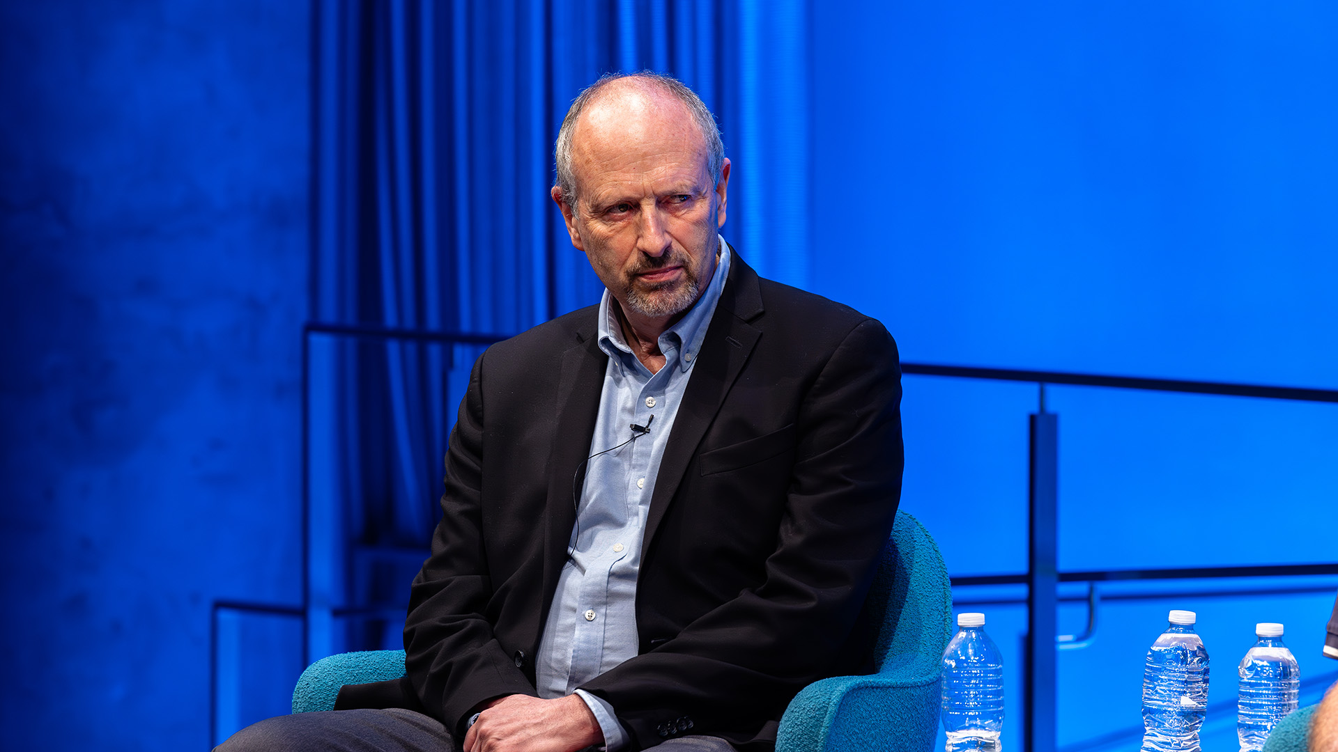 A single speaker sitting on stage, in front of a blue backdrop.