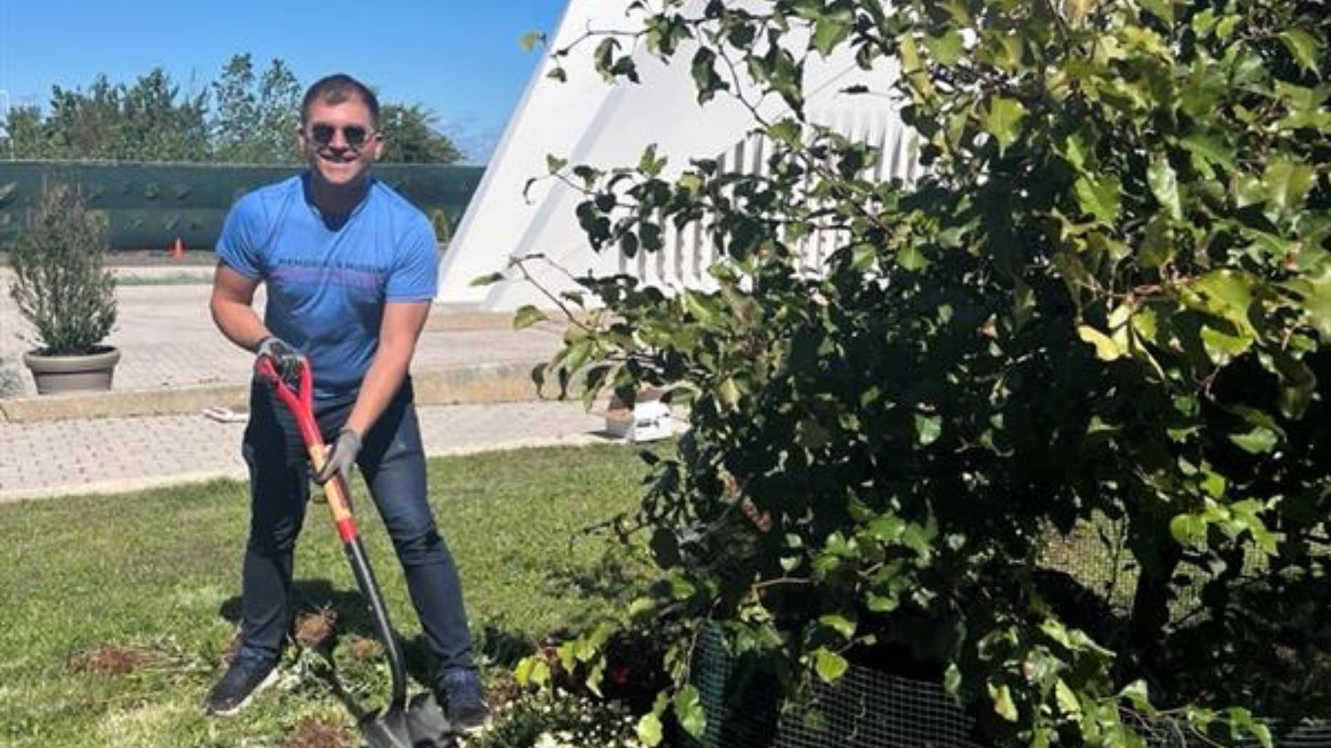 A Visionary smiles while holding a gardening shovel near a tree