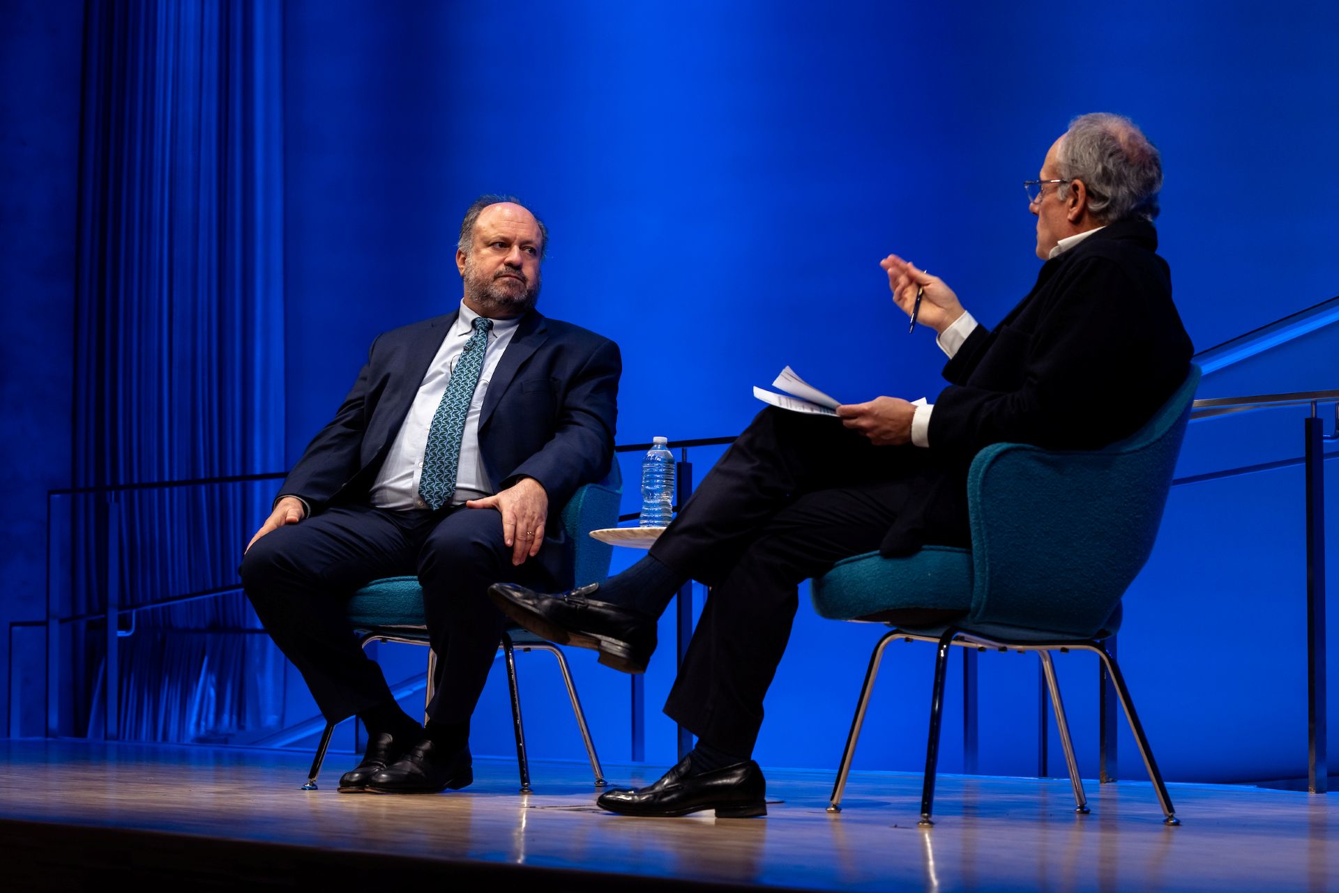 Bernard Haykel and Cliff Channing speak together on stage. Cliff is gesturing and Bernie is listening. A blue background is visible.