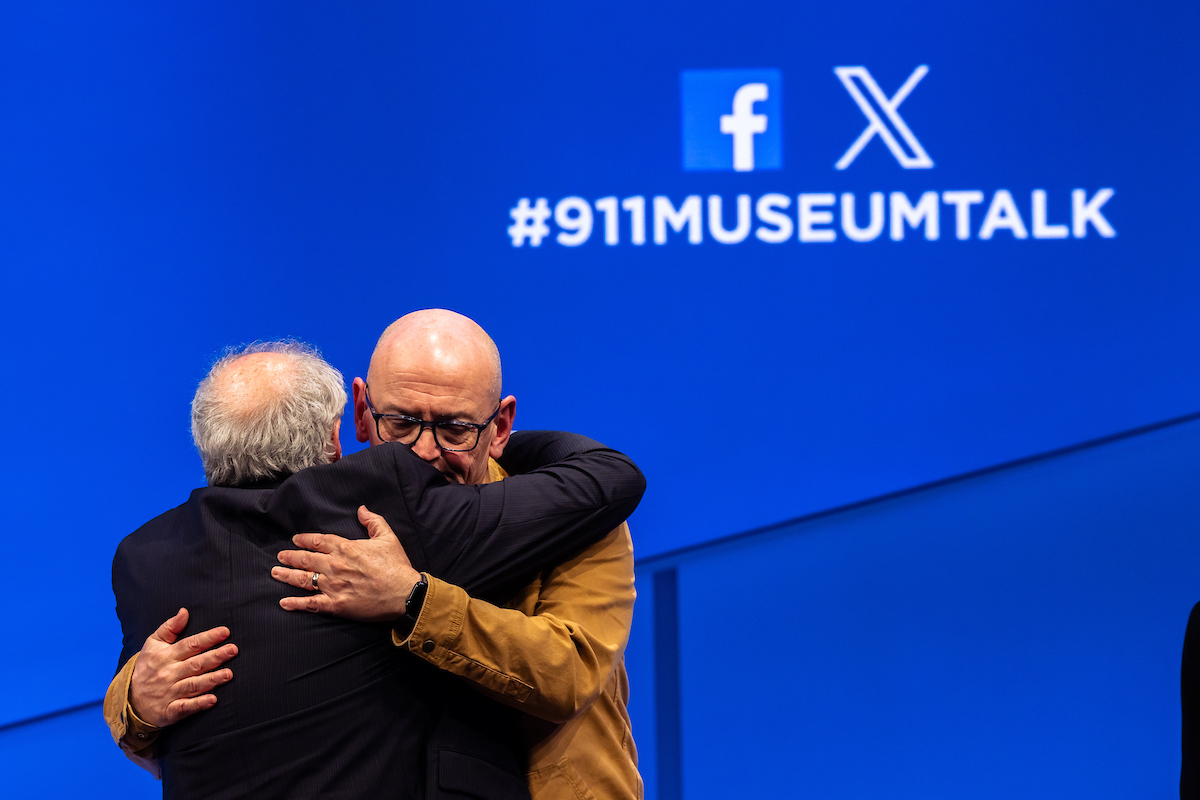 Two men hug on the stage in front of a blue screen or banner