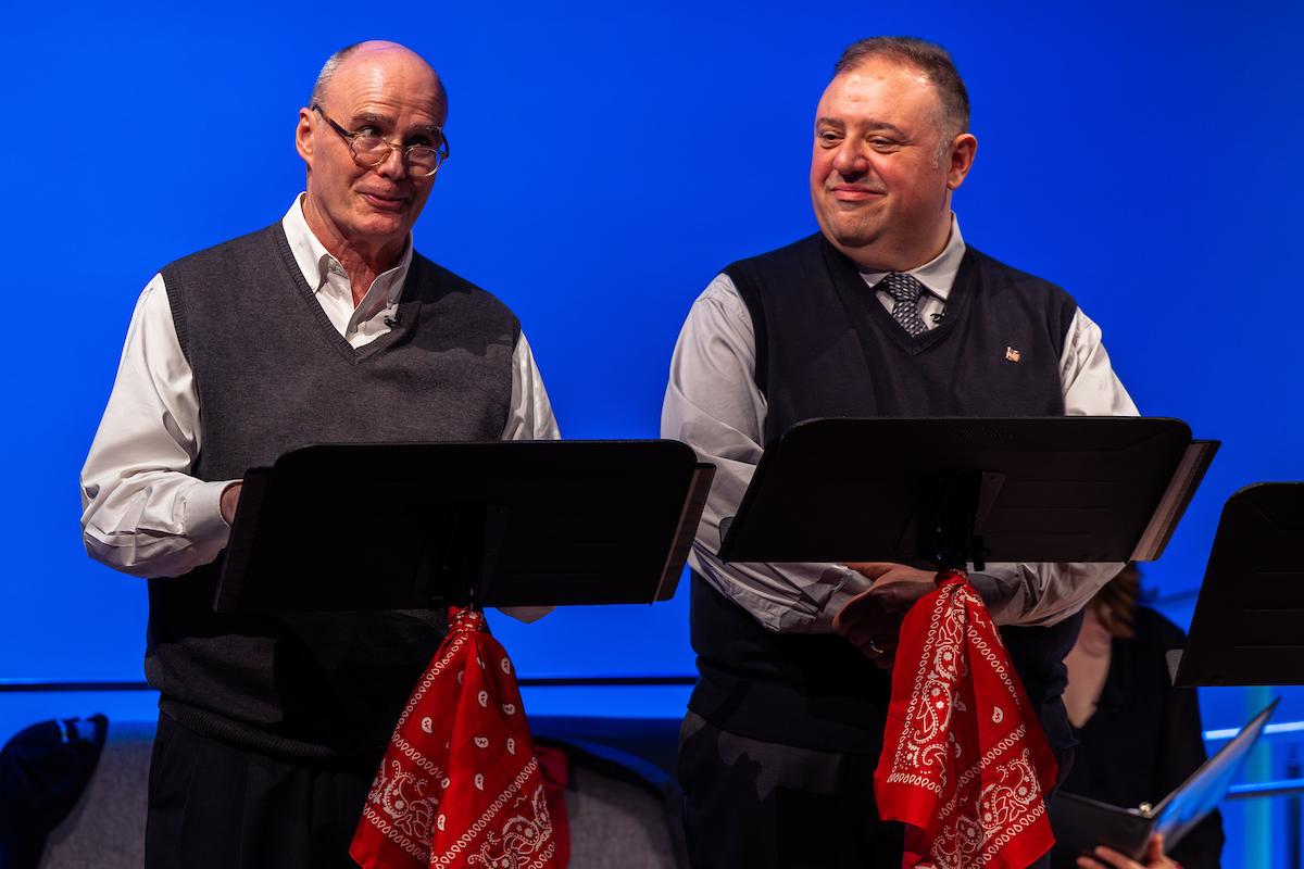 two men read from scripts on stands against a blue background