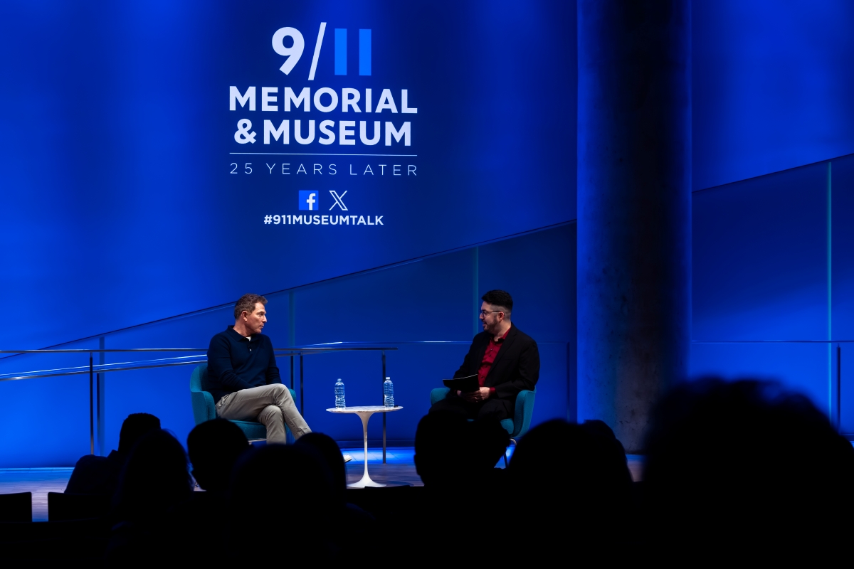 Two men sit on a stage with 911 Memorial Logo behind them 