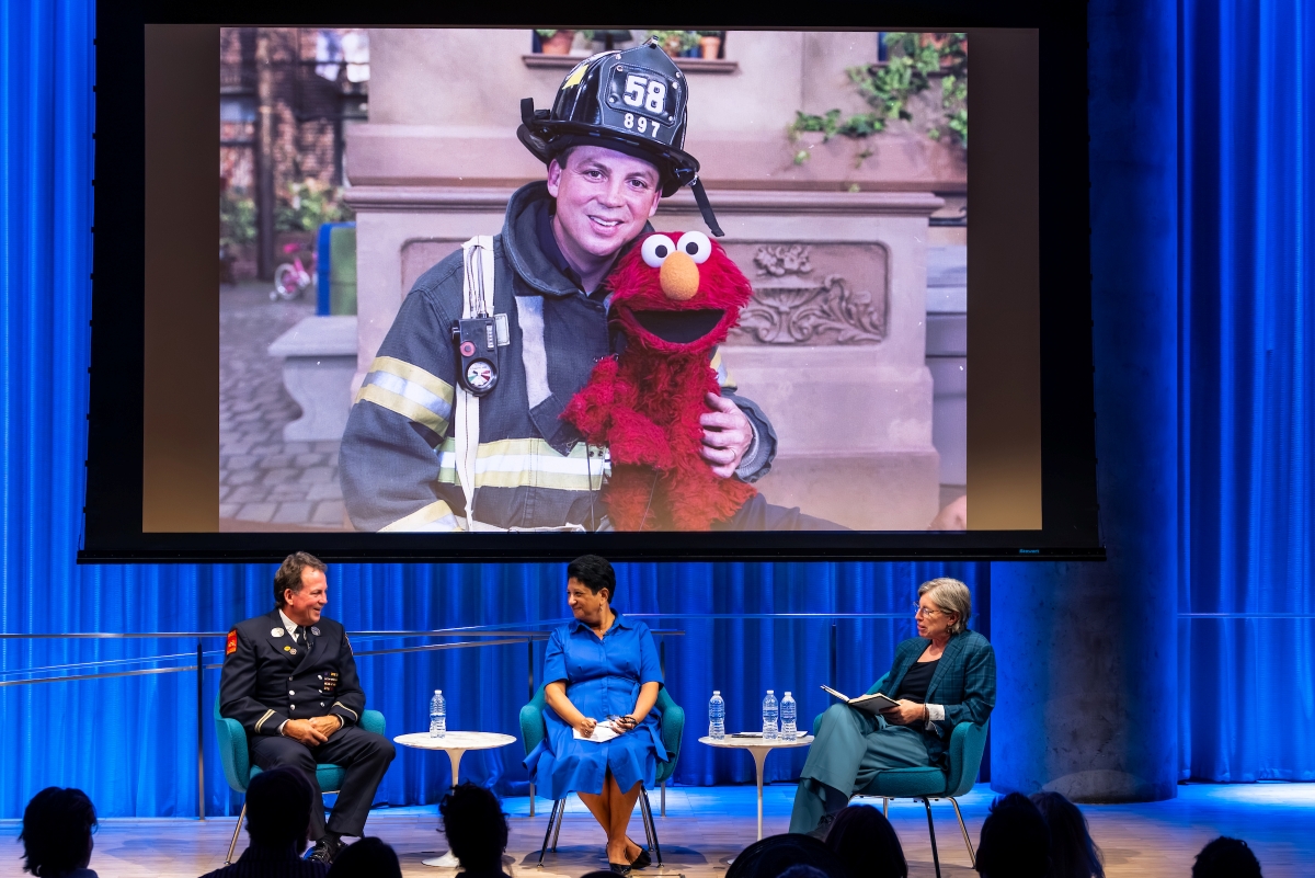 Three people on stage sit under a large screen showing a photo of the man on the left from years before when he and Elmo from Sesame Street were posed together. The man and red muppet embrace and the man is wearing his full fire fighter uniform including hat.