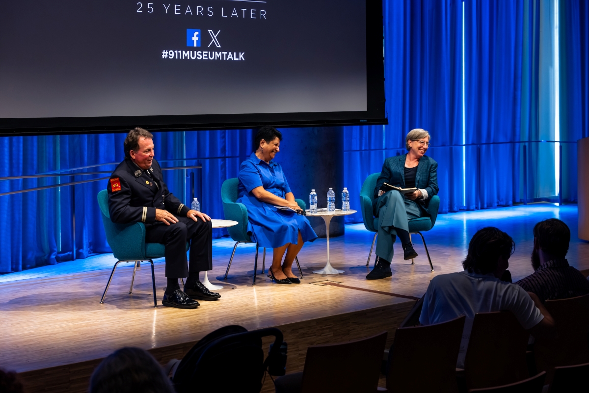 Three people sit on stage with the 9/11 Memorial & Museum log visible on a screen behind them.
