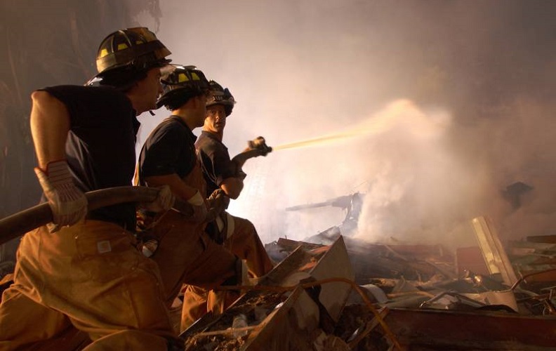 A historical photo from Ground Zero shows three firefighters hosing down a smoky pile of debris at night. A flood light illuminates the smoke and partially silhouettes the three firefighters. 