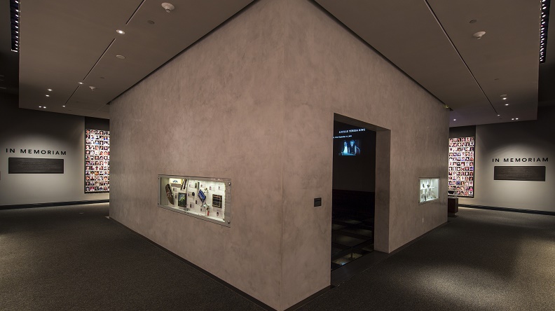 An interior look at the In Memoriam exhibition. Personal artifacts that belonged to victims are displayed on the walls of an inner chamber in which their profiles are projected. Some of the 2,983 portrait photographs of victims are on walls to the left and right of the chamber. 