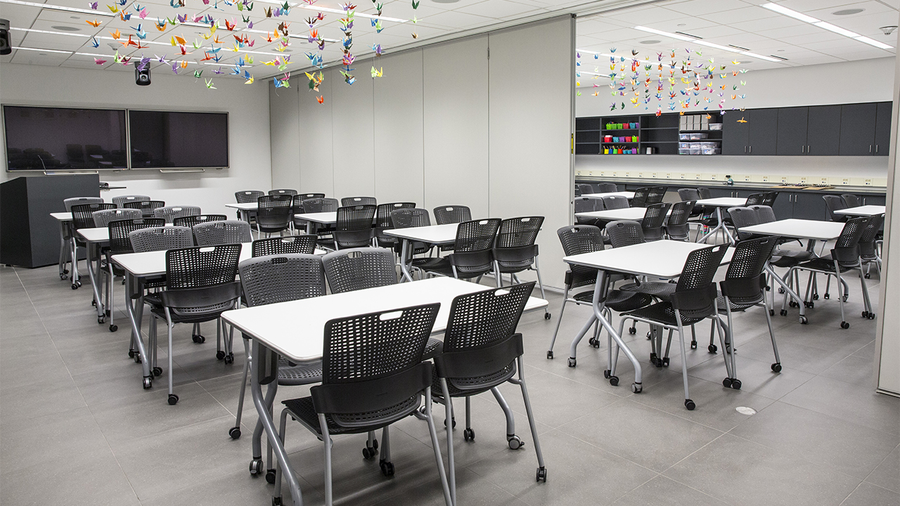 Black chairs and white tables fill an empty Education Center. There are two classrooms separated by a room divider. Colorful origami birds hang over the tables. Presentation screens and a lectern are at the front of the room.