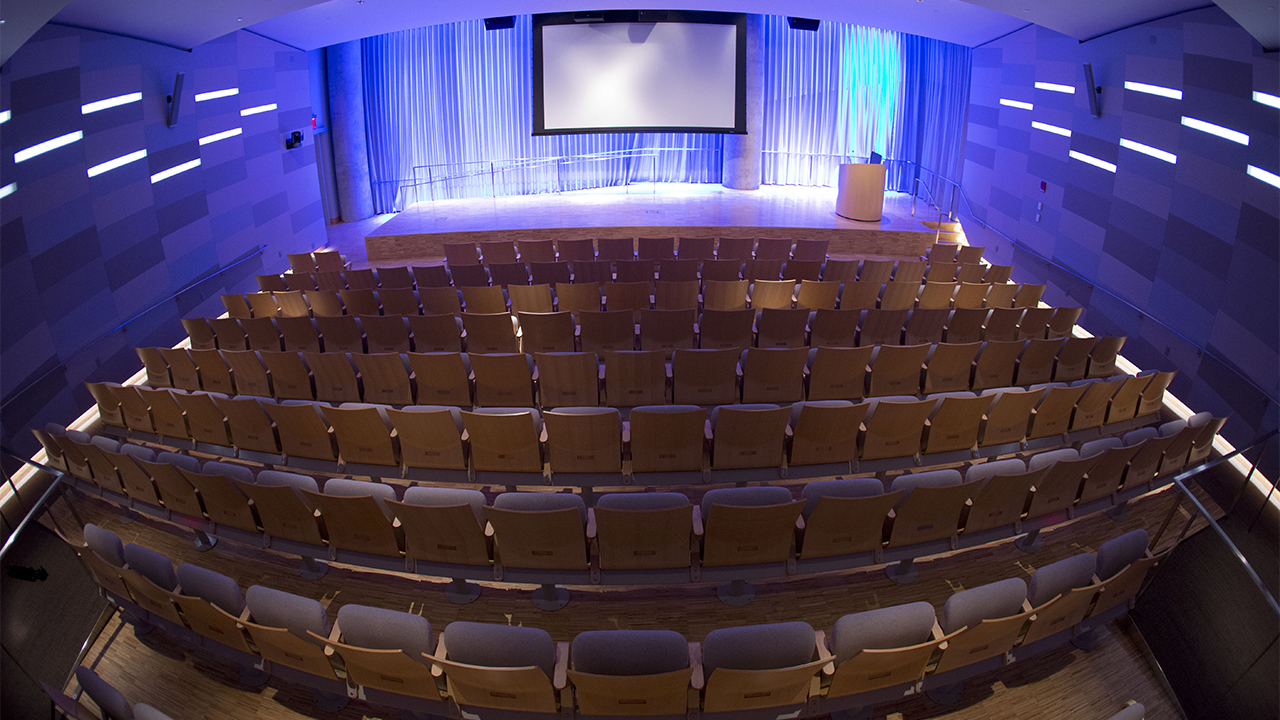 An empty Museum Auditorium is seen from above. Wooden theater seats are in the foreground. Further back, lights illuminate a stage, which has a lectern and lowered video screen.