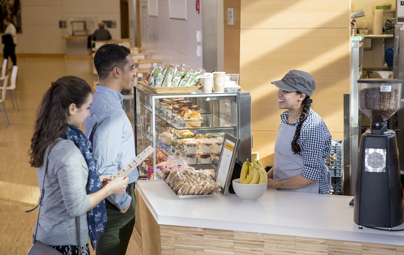 A woman behind a register greets a man and woman at the Pavilion Cafe. The two customers are looking through a menu as they speak with the cashier. Fruit, pastries and other food are in a display case.