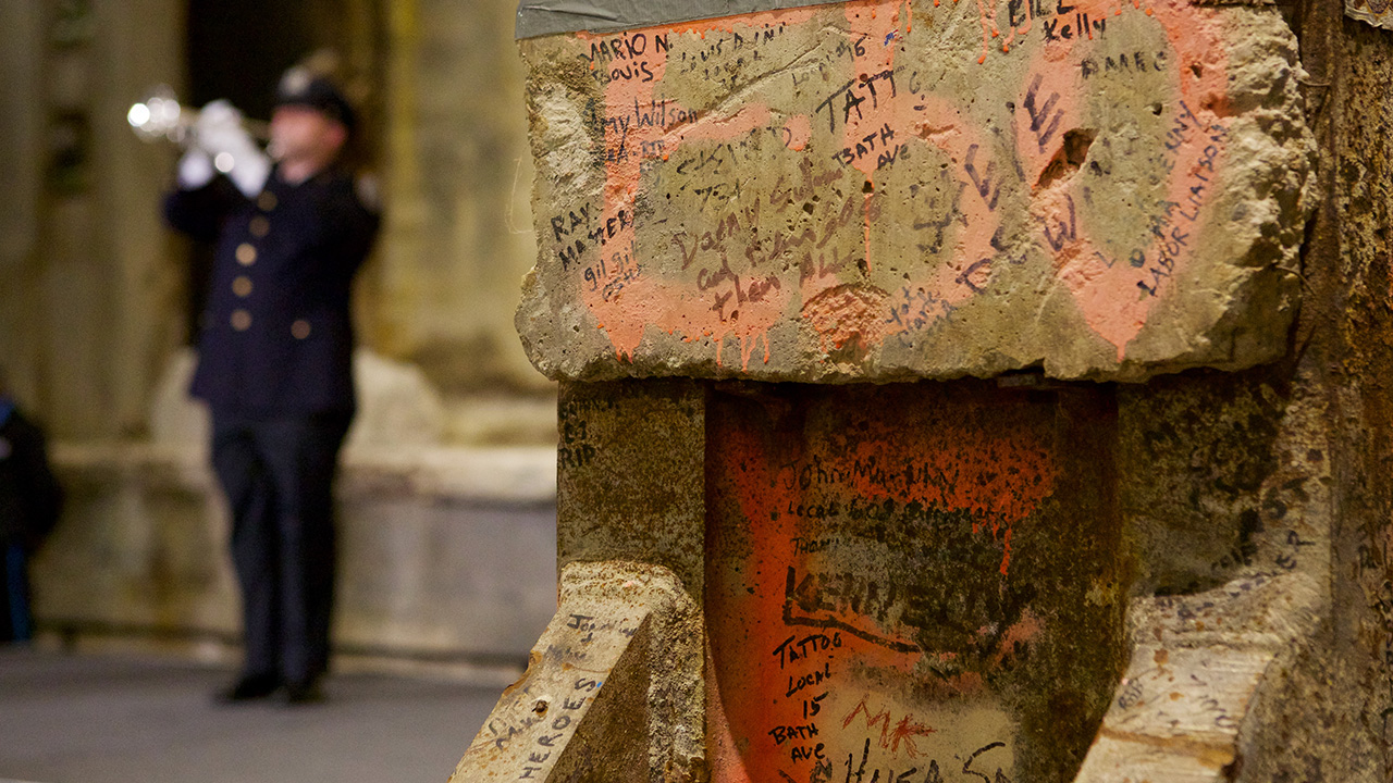 An NYPD officer in formal clothing plays “Taps” on a trumpet near the Last Column during a ceremony in Foundation Hall. The officer is to the left and out of focus. In the foreground, details of the Last Column, including handwritten messages and orange marking paint, are visible.