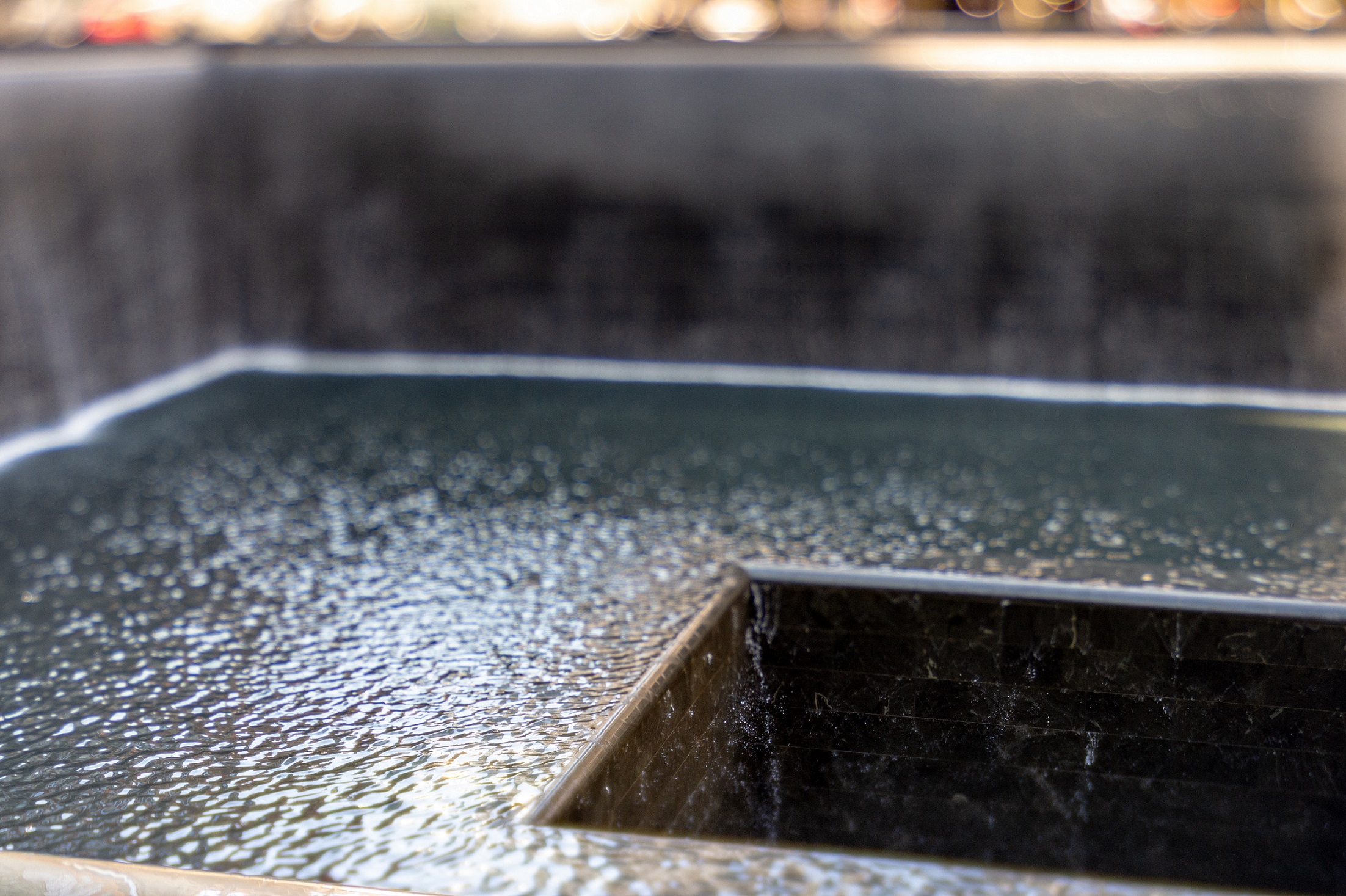 Water flows down two walls and disappears into a void at the center of a Memorial reflecting pool. Sunlight creates shadows on the pooled water. 