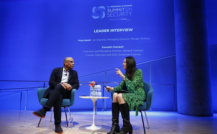 A man in a suit and a woman in a green dress sit in chairs onstage at the Summit on Security. A projected outline of the event behind them says the event they’re taking part in is about “identifying new vulnerabilities.”