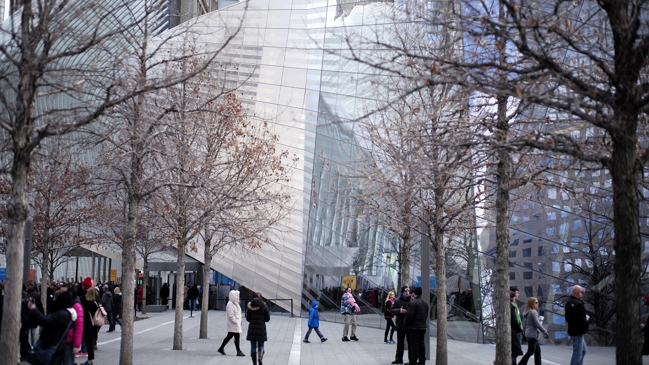 Visitors walk around the 9/11 Memorial plaza in front of the Museum's facade on a winter day. The trees are bare. 