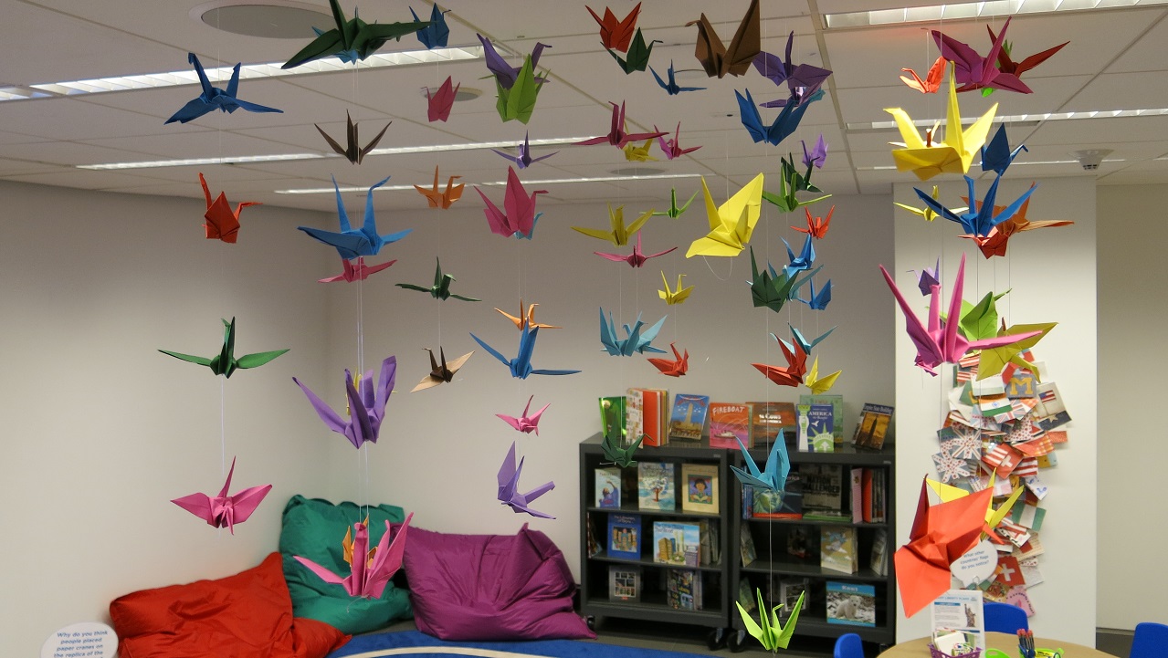 Paper origami cranes of many colors hang from a classroom ceiling in many chains. In the background of the photo is a bookcase and several brightly colored pillows propped against the classroom wall. 