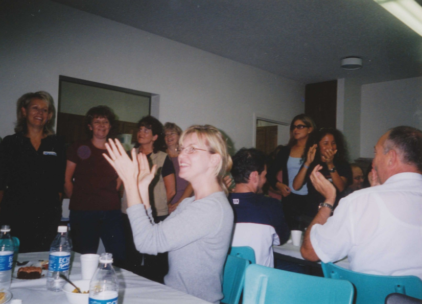 Photograph taken by Jackie Pinto of several people inside the dining room of a church. Back of original photograph has a handwritten inscription: "appreciation applause for local people in dining room." 