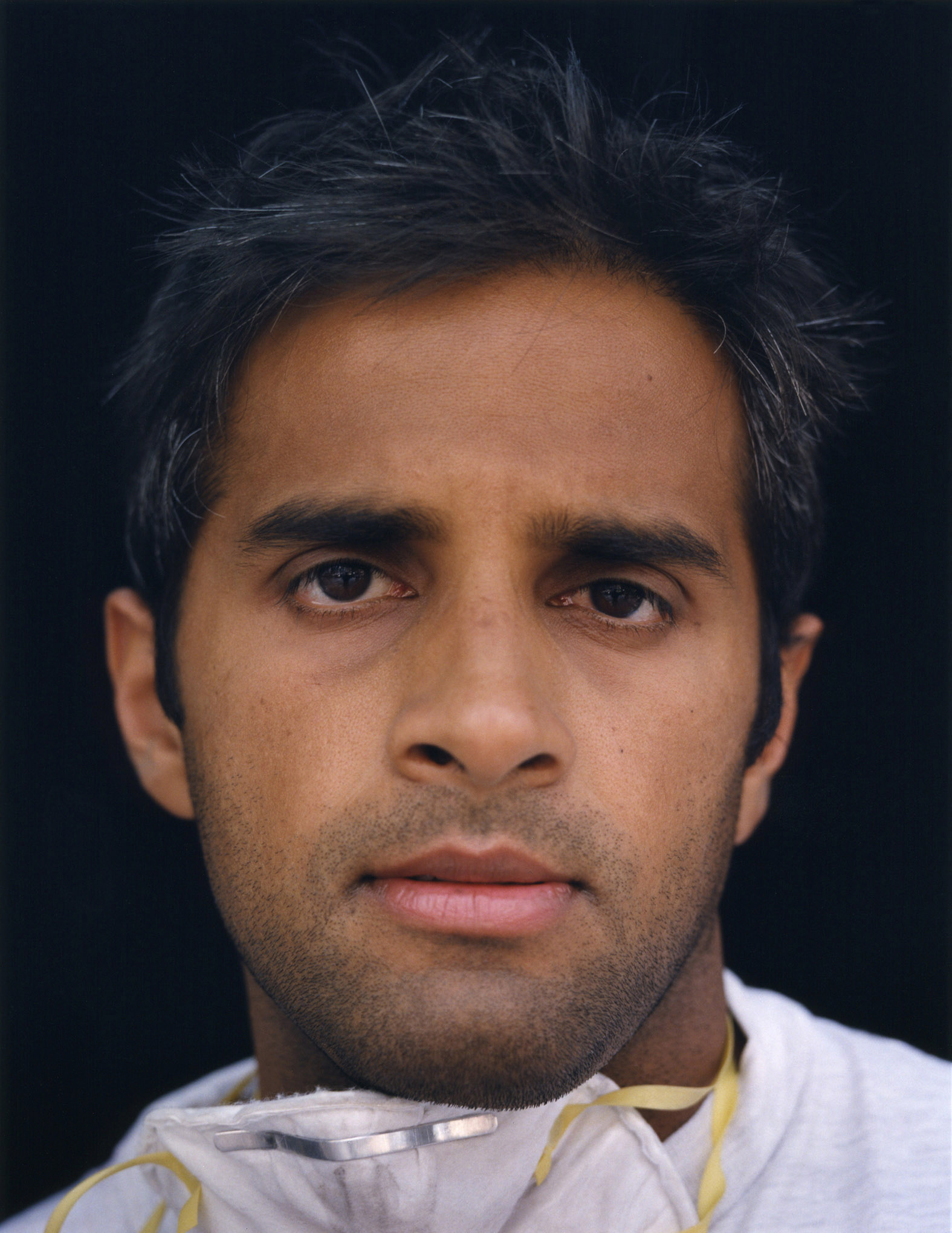 A man with a face mask around his neck stares directly ahead.  He  looks tired but calm. 