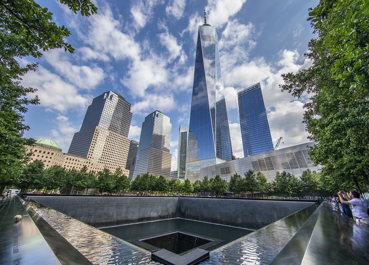A view of the 9/11 Memorial on a partly cloudy day. Clouds are reflected in the facade of One World Trade.