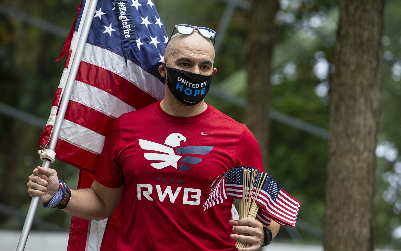 A man waving an American flag and wearing a T-shirt that reads "Team Red White & Blue" wears a face mask and is about to hand out small American flags. 