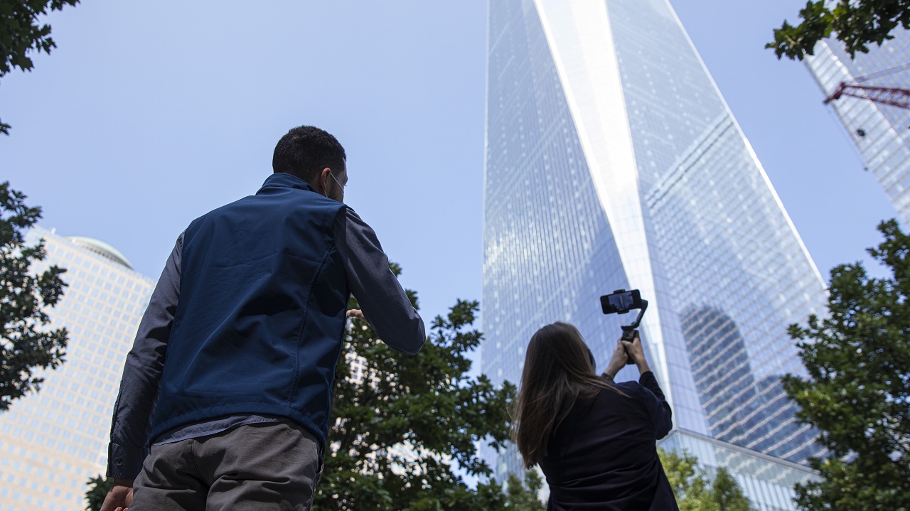 A woman with a smartphone films One World Trade while a tour guide speaks at stands behind her on the 9/11 Memorial plaza.