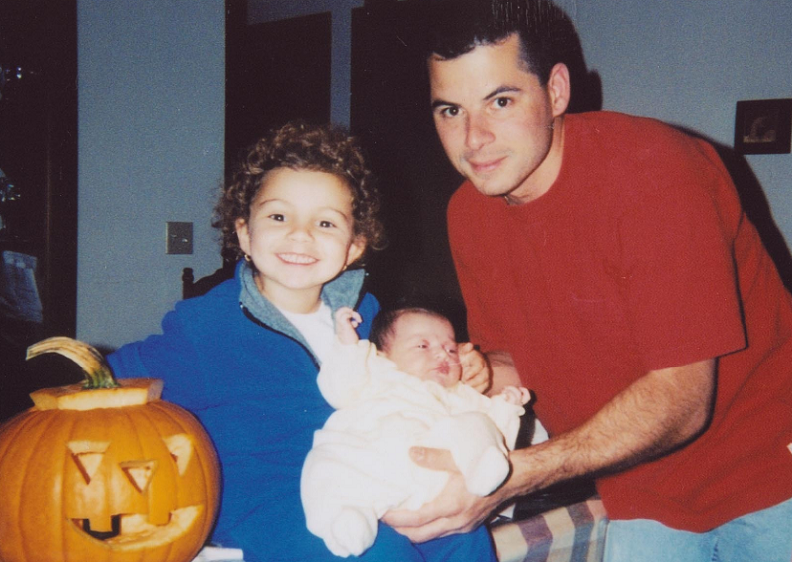 A father holds his infant  child and stands next to his kindergarten-aged daughter, who smiles brightly next to a carved pumpkin. 