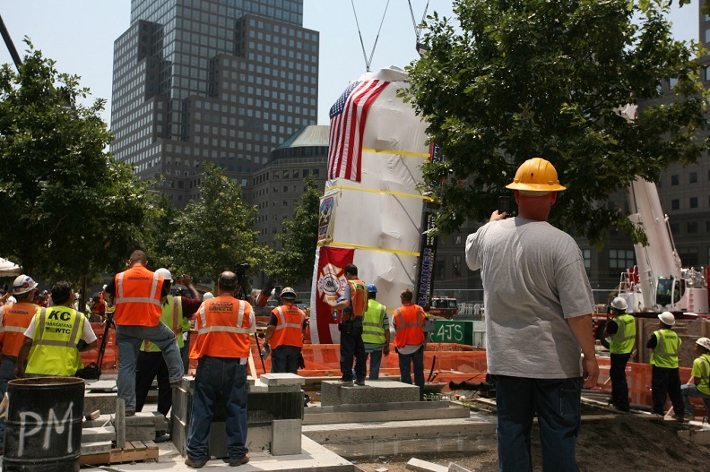 A firetruck covered in a white tarp and American flag is lowered into the 9/11 Memorial Museum through a hole in the 9/11 Memorial plaza. Men in hardhats supervise the lowering. 