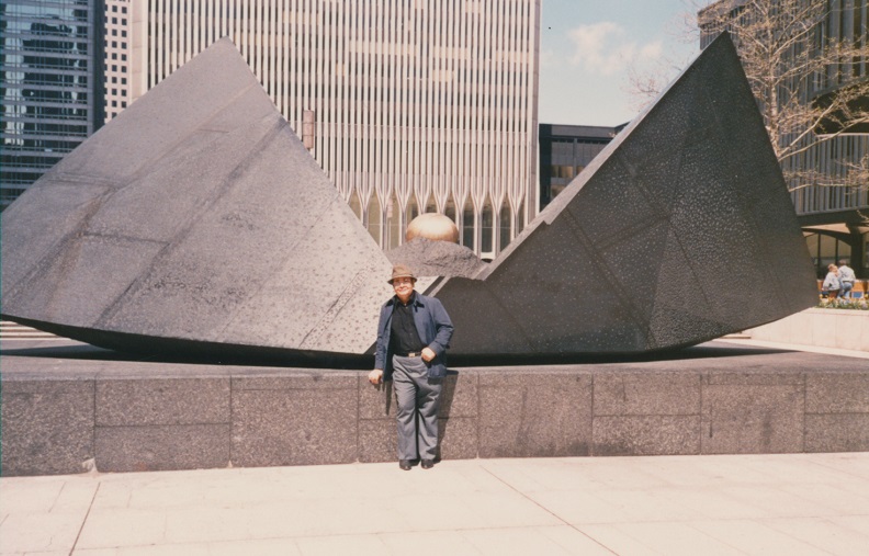 A man stands in front of an abstract granite sculpture of two pyramids attached on the base and tilted upwards. The ground level of a Twin Tower is visible in the background. 