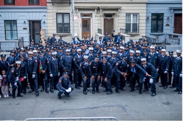 Several rows of uniformed firefighters stand in front of three urban doorways painted, from left to right, light blue, beige, and light blue 