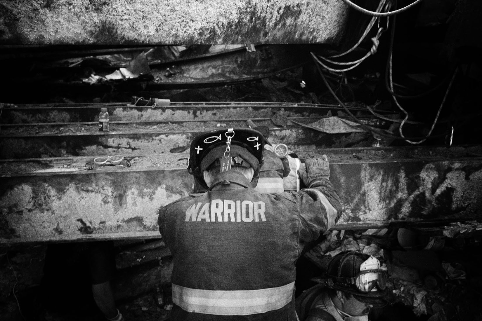 Black and white photo of a firefighter at Ground Zero, back to the camera. The back of jacket says Warrior, and Christian fish and crosses are drawn on helmet brim.  